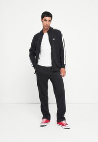 Black zip-up jacket with white stripes, paired with black sweatpants. Model wears a white t-shirt and red sneakers. Smooth fabric texture.