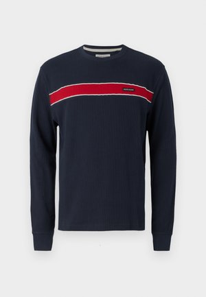 Mornarska dolga majica z rebrastim vzorcem, ki ima vodoravno rdečo črto z belimi poudarki in logo na prsih. Okrugli izrez z prilegajočimi man�šetami.