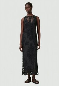 Vestido maxi preto sem mangas com padrões florais bordados, corpete transparente e bainha de renda recortada. Usado com sandálias pretas.