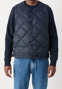 Dunkelblau gesteppte Puffer-Weste mit rundem Ausschnitt, Druckknöpfen und Logodetail. Getragen über einem dunkelblauen Sweatshirt mit blauen Jeans.