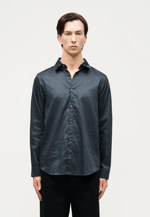 Άνδρας που φοράει σκούρο πουκάμισο με μοτίβο και κουμπιά, μακρυμάνικο, στέκεται μπροστά σε απλό ανοιχτό φόντο.