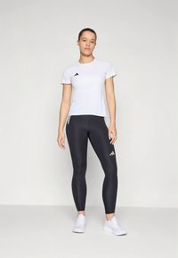 Balta īsroku t-krekls ar melnu logo, kombinēts ar melnām legingām ar akcentētu logo. Modelis valkā baltas sporta kurpes.