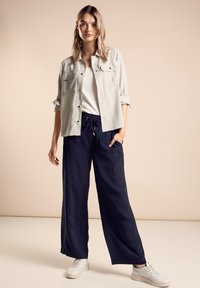 Vaaleanharmaa nappipaita valkoisen t-paidan päällä, yhdistettynä tummansinihin leveälahkeisiin housuihin ja valkoisiin lenkkareihin. Silkkinen kangas, rento malli, epämuodollinen muotoilu.