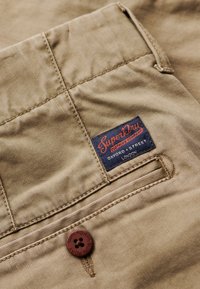 Khaki stof met een gladde textuur, voorzien van een blauw label met "SuperDry" branding en een bruine knop op de zak.