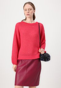 Rosa Strickpullover mit weitem Ausschnitt und lockeren Ärmeln, kombiniert mit einem figuroptimierenden bordeauxfarbenen Lederrock und einer kleinen schwarzen Handtasche.