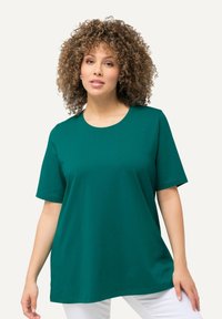 T-shirt in cotone color turchese con scollo rotondo e maniche corte, caratterizzato da una vestibilità morbida e una texture liscia. Il modello indossa pantaloni bianchi.
