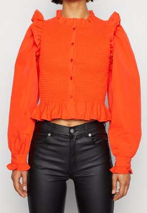 Red smocked crop top with ruffled neckline and sleeves, featuring a button front and a flared hem. Paired with black high-waisted pants.