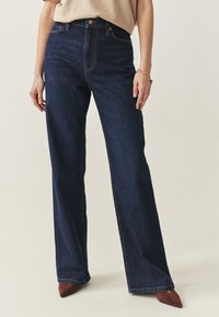 Blugi largi, cu talie înaltă, de culoare albastru închis, cu design clasic cu cinci buzunare și cusături contrastante. Purtați cu pantofi cu toc maro și vârful ascuțit.