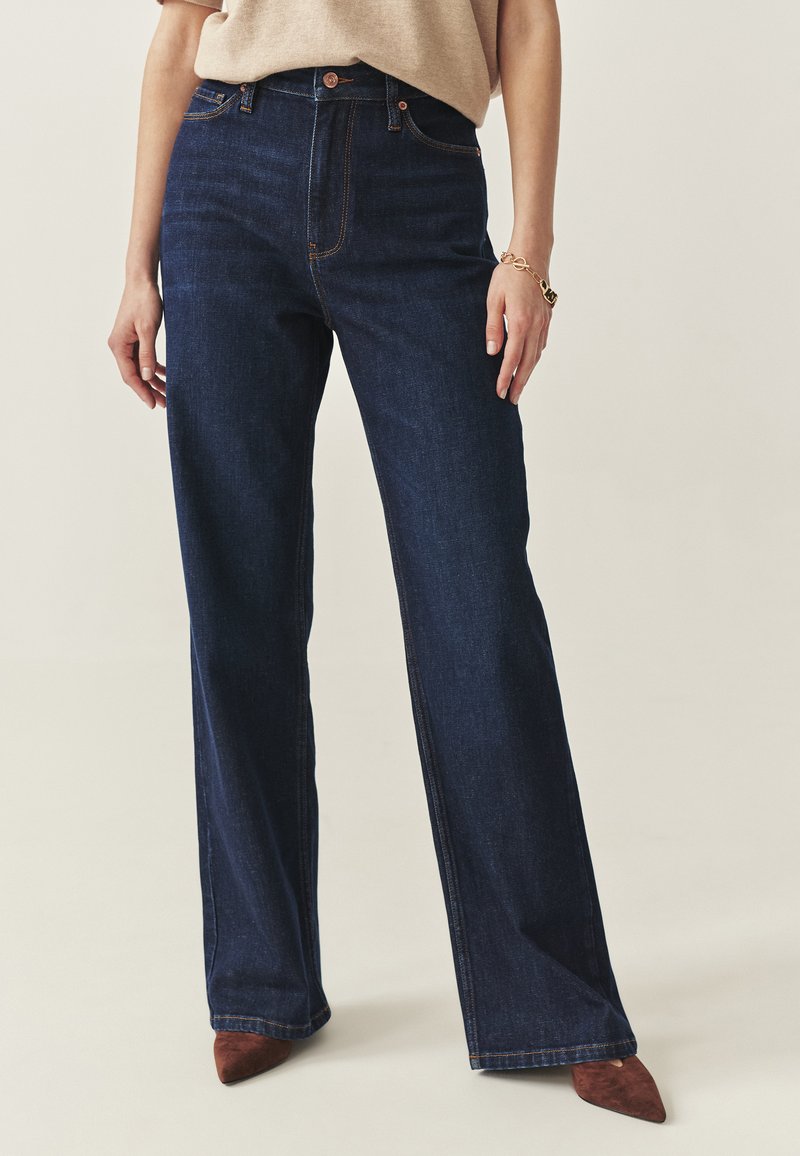 Blugi largi, cu talie înaltă, de culoare albastru închis, cu design clasic cu cinci buzunare și cusături contrastante. Purtați cu pantofi cu toc maro și vârful ascuțit.