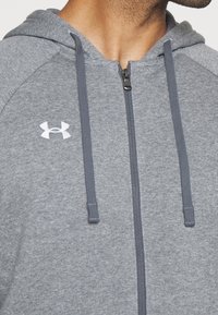 Sudadera con cremallera gris hecha de un material suave y texturizado. Presenta un logo blanco, cordones ajustables y un cierre con cremallera.