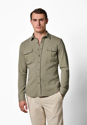 Mann trägt ein olivgrünes Hemd mit Knöpfen und zwei Brusttaschen sowie beige Hose, steht vor einem schlichten weißen Hintergrund.