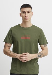 Olivegrønn bomull T-skjorte med en rød rektangulær grafikk med "A.R.C." og japanske tegn. Korte ermer, klassisk rund hals.