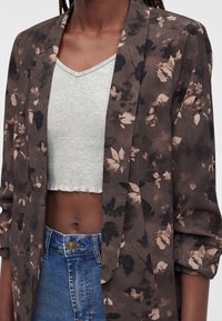 Gros plan d'une personne portant un blazer marron à motifs floraux sur un haut court gris clair et un jean bleu taille haute.