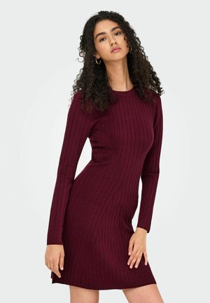 Femme avec des cheveux bouclés portant une robe moulante à manches longues en maroon côtelé, se tenant avec une main reposant sur sa cuisse contre un fond blanc.