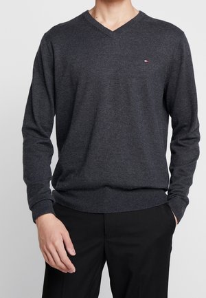 Mann trägt dunkelgrauen V-Ausschnitt-Pullover mit kleinem Logo auf der Brust und schwarze Hose, steht mit einer Hand in der Tasche.