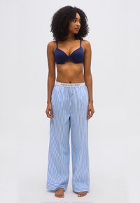 Γυναίκα με σγουρά μαλλιά που φοράει μπλε σουτιέν και άνετο παντελόνι πιτζάμας με μπλε και άσπρες ρίγες και ζώνη Polo Ralph Lauren, όρθια ξυπόλητη και κοιτάζει μπροστά.