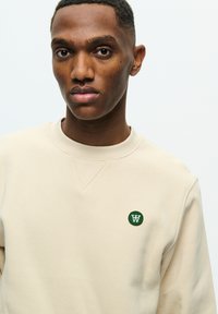 Beige sweatshirt featuring a round green logo with a white "W" on the left chest, crew neck, and a textured, soft fabric.
