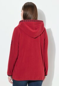 Punainen fleece-takki hupulla, jossa on kuvioitu trimmaus kauluksessa ja tummansinisiä yksityiskohtia hihoissa. Pehmeä rakenne ja rento istuvuus.
