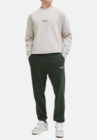 Helles beige Sweatshirt mit "JJ Studios"-Logo auf der Brust, kombiniert mit dunkelgrünen Jogginghosen mit Logo an der Seite, getragen mit weißen Sneakers.