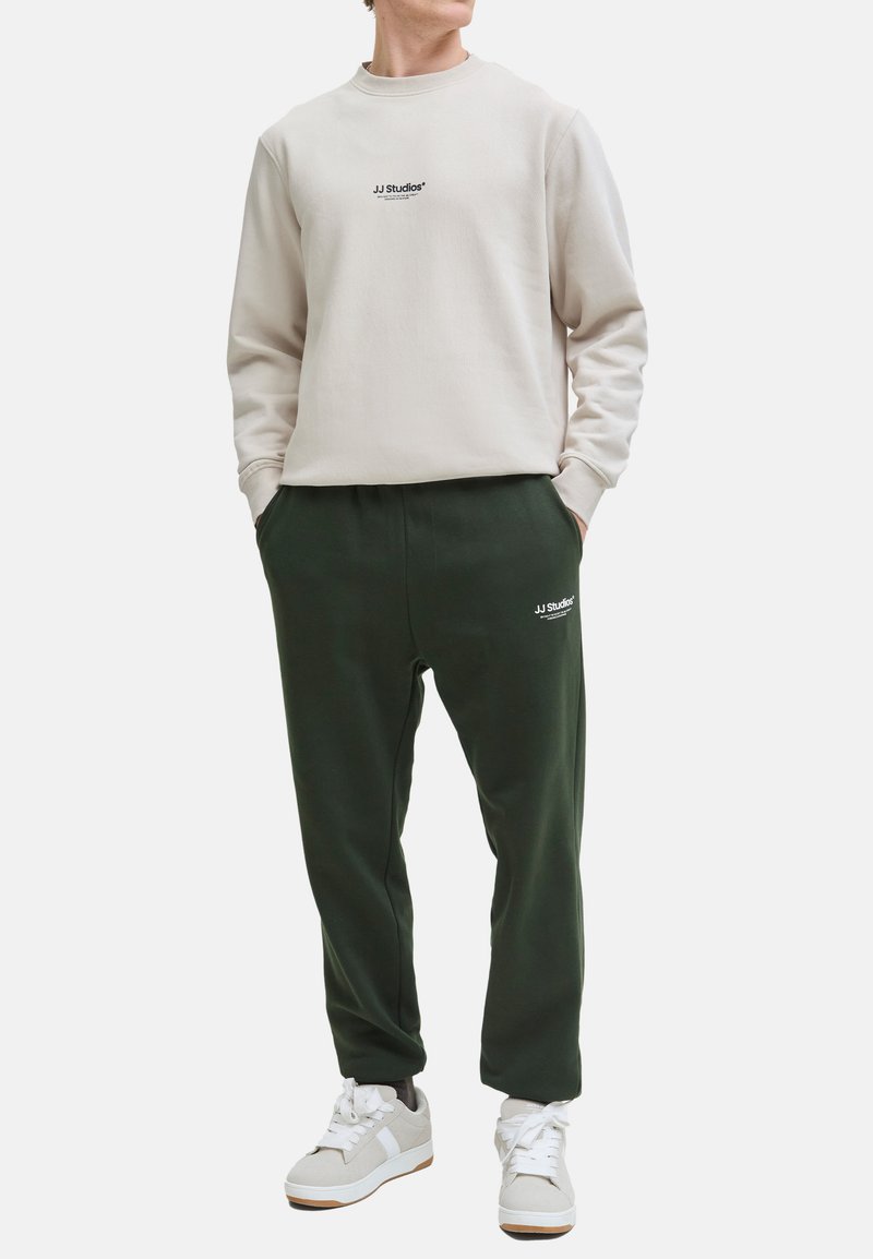 Helles beige Sweatshirt mit "JJ Studios"-Logo auf der Brust, kombiniert mit dunkelgrünen Jogginghosen mit Logo an der Seite, getragen mit weißen Sneakers.