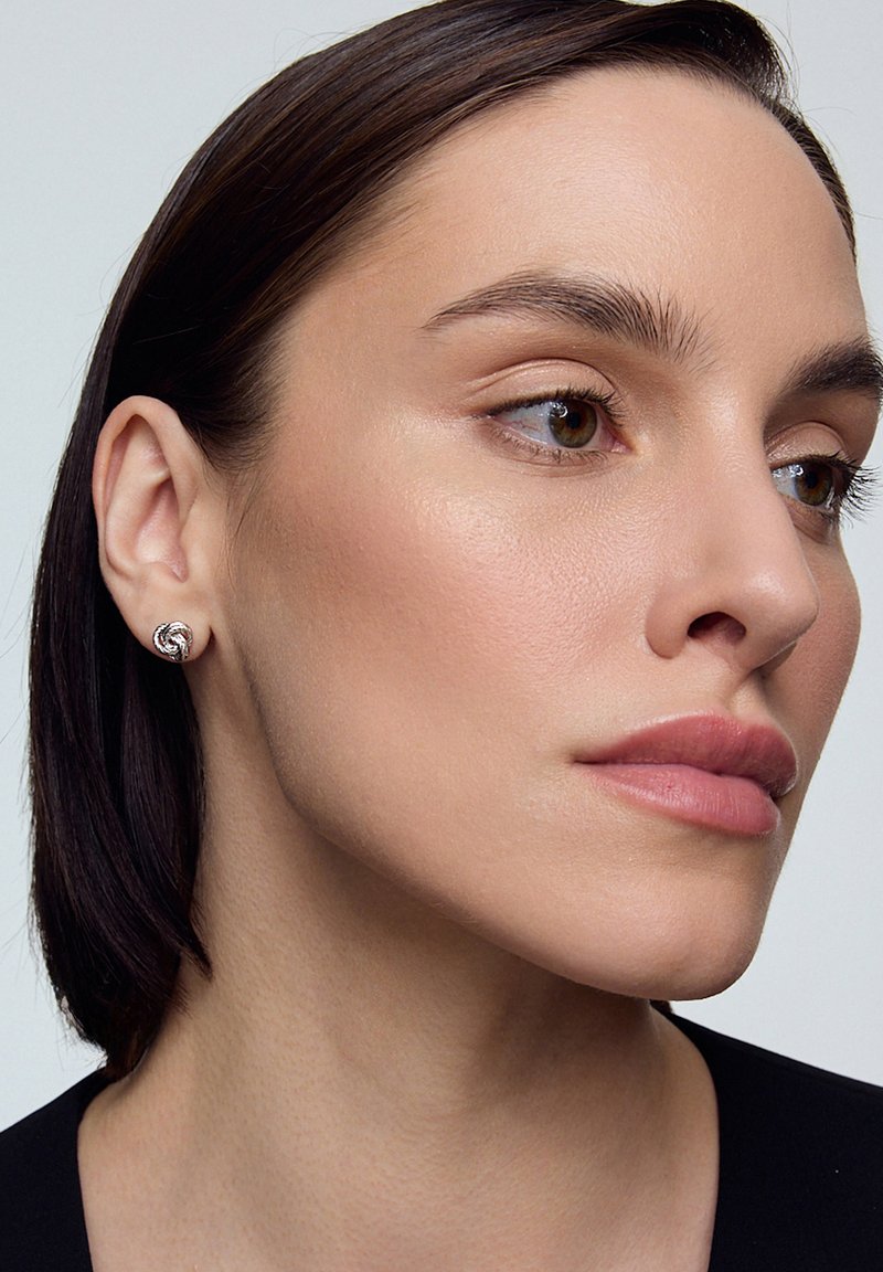 Srebrne kolczyki z ćwiekami o zakręconym wzorze, noszone na uchu modelki, podkreślone gładką skórą i neutralnym makijażem.