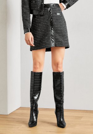 Gonna in tweed nera con dettagli in pelle, caratterizzata da bottoni frontali e una forma svasata. Abbinata a stivali neri in pelle con stampa coccodrillo alti fino al ginocchio.