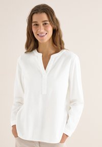 Jeune femme souriante, portant un chemisier blanc texturé à manches longues avec un col en V et les mains dans les poches, sur un fond neutre.