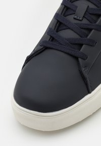 Baskets en cuir bleu marine avec bout rond, semelle en caoutchouc blanc et lacets plats. Présente une texture lisse et un design minimaliste.