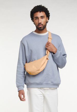 Peachfarvet bæltetaske med en glat, mat tekstur, båret over kroppen. Kombineret med en lyseblå sweatshirt og hvide bukser.