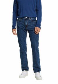 Hombre vestido con un suéter azul, jeans azul oscuro y zapatillas blancas, de pie frente a un fondo blanco.