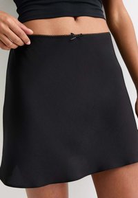 Minifalda negra hecha de tela ligera. Presenta un borde con volantes y un pequeño lazo decorativo en la cintura. Textura suave.