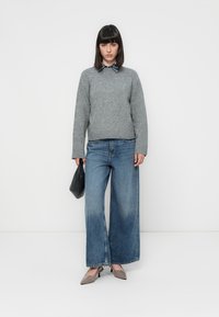 Maglione grigio lavorato a maglia, ampissimi jeans in denim blu e décolleté grigie a punta. Regge una borsa nera, in piedi contro uno sfondo semplice.