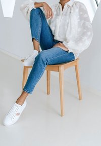 Blouse blanche à motif floral texturé, jean bleu et baskets blanches avec des accents roses, assise sur une chaise en bois clair devant un fond neutre.