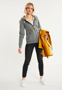 Sudadera gris con cremallera y capucha forrada, detalles de cremallera amarillos, combinada con pantalones ajustados negros y zapatillas blancas, sosteniendo una chaqueta amarilla.