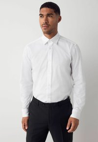 Weißes Langarmhemd mit einem dezenten geometrischen Muster, klassischem Kragen und Knopfverschluss. Kombiniert mit schwarzen Hosen.