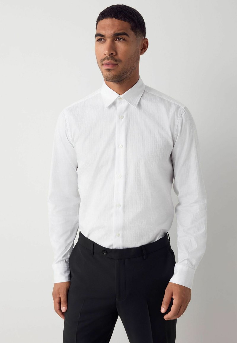 Weißes Langarmhemd mit einem dezenten geometrischen Muster, klassischem Kragen und Knopfverschluss. Kombiniert mit schwarzen Hosen.