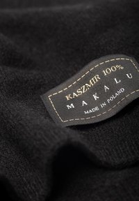 Czarna tkanina kaszmirowa z widoczną metką mówiącą "100% KASZMI R MAKALU WYPRODUKOWANE W POLSCE." Charakteryzuje się delikatną teksturą i haftowanymi detalami na metce.