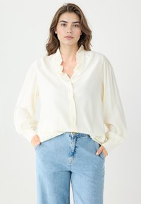 Kremowa bluzka z falbaną przy kołnierzyku, z przodem zapinanym na guziki i długimi, bufiastymi rękawami. W zestawie z jasnoniebieskimi dżinsami.