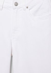 Blugi albi din denim, prim-plan, care arată nasturele frontal, zona fermoarului și buzunarul frontal drept cu nit.