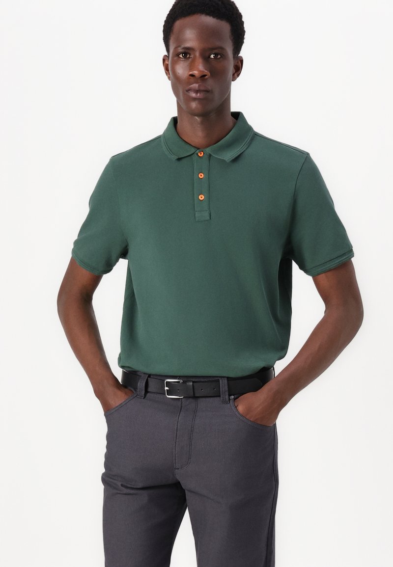 Groene polo gemaakt van katoen met een gladde textuur, voorzien van een oranje knoopsluiting. Gekocht met donkergrijze broeken en een zwarte riem.