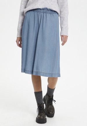 Femme portant une jupe bleu clair jusqu'aux genoux, un pull gris à motifs, des chaussettes grises côtelées et des bottes noires épaisses à lacets, debout sur un fond blanc.