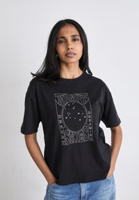 Juodas per didelis t-shirt, pagamintas iš medvilnės, su baltu tekstu ir žvaigždžių raštu, sakantis "Naktiniai dangūs dykumos sapnai", centrinėje dalyje priekyje.