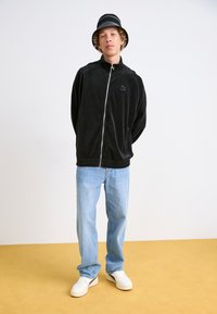 Svart fløyelsjakke med glidelås og ståkrage, kombinert med lyseblå jeans og hvite sneakers. Sort-hvite stripete bøttehatt.