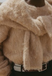 Beige, oversized sweater with a fuzzy texture and a loose front tie. Worn over dark clothing, complemented by a contrasting belt.