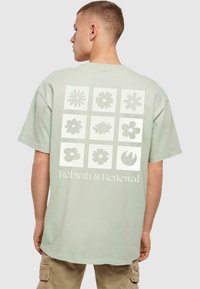 Lys grøn bomulds t-shirt med et netværk af blomsterdesigns og teksten "Genfødsel & Fornyelse" i hvid på ryggen. Korte ærmer, afslappet pasform.
