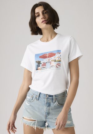 Femme aux cheveux courts et foncés, portant un t-shirt blanc avec une scène de plage et un short en jean bleu clair déchiré, sur un fond uni.