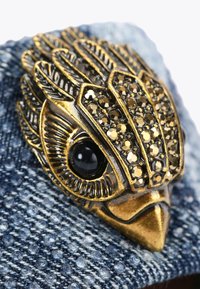 Material denim con un design di gufo in tonalità oro; presenta gemme nere per gli occhi e dettagli metallici intricati sulle piume, superficie texturizzata.