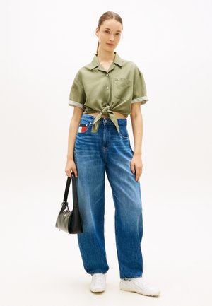Mladá žena v olivovo zelenej viazanej košeli, modrých džínsach s vysokým pásom a logom, biele tenisky, drží čiernu kabelku.