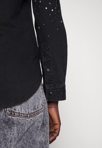 Black denim shirt with a curved hem and long sleeves adorned with scattered rhinestones, featuring buttoned cuffs and a textured fabric.