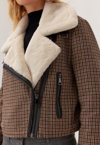 Brązowa i czarna kurtka w desenie houndstooth z dużym kremowym sztucznym futrem wokół kołnierza, z zamkiem z przodu i boczną kieszenią na zamek, noszona na czarnym prążkowanym topie.